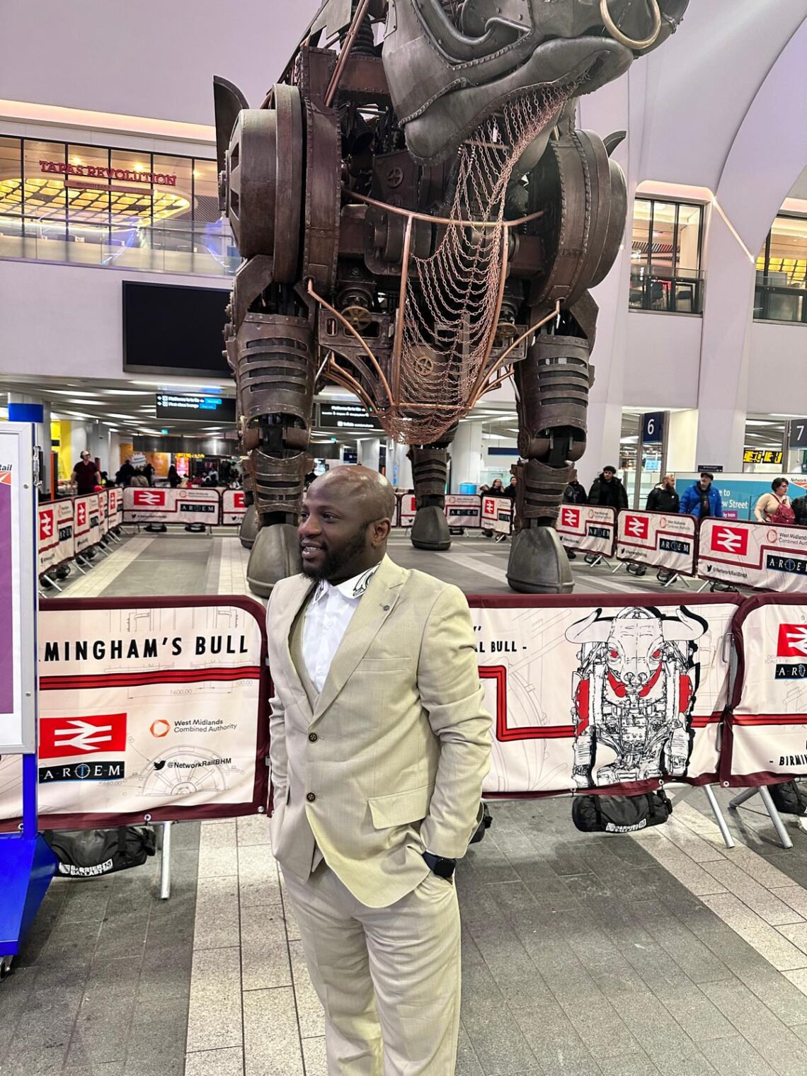 Taiwo Standing in front of the bull statue in Birmingham wearing a butter colored suit and a white shirt with design on its lapel.